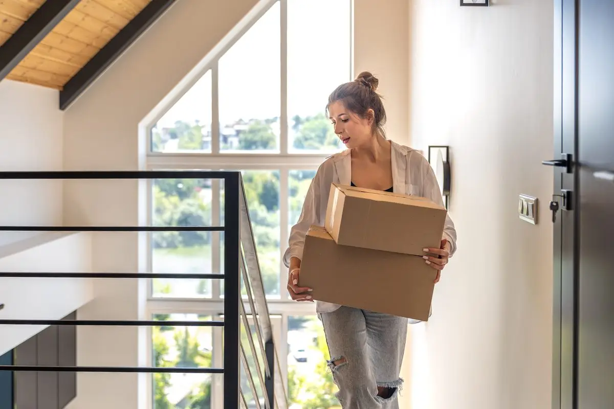 A woman carrying two cardboard boxes through a bright hallway