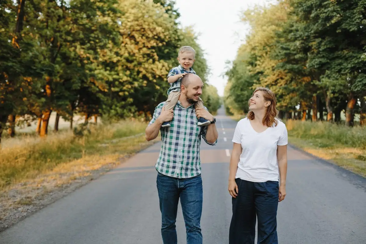A father carrying his young son on his shoulders while the mother walks beside them on a tree-lined road