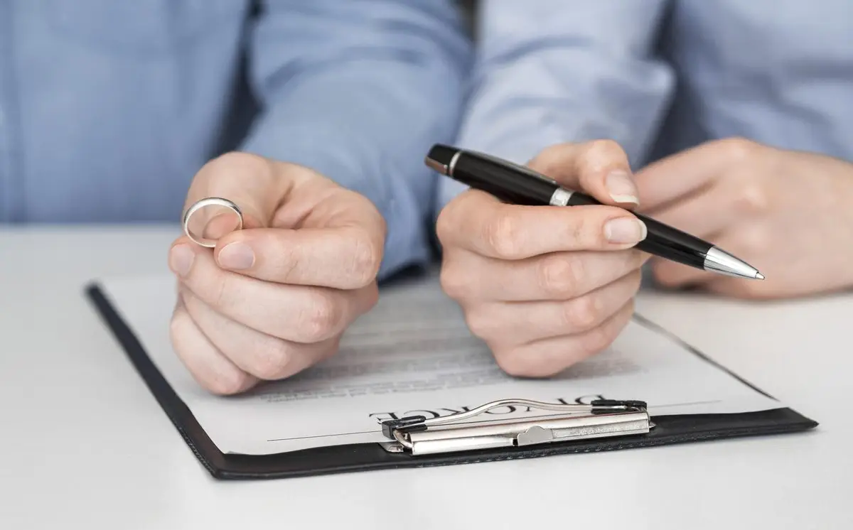 A hand holding a wedding ring beside a pen and a document on a clipboard
