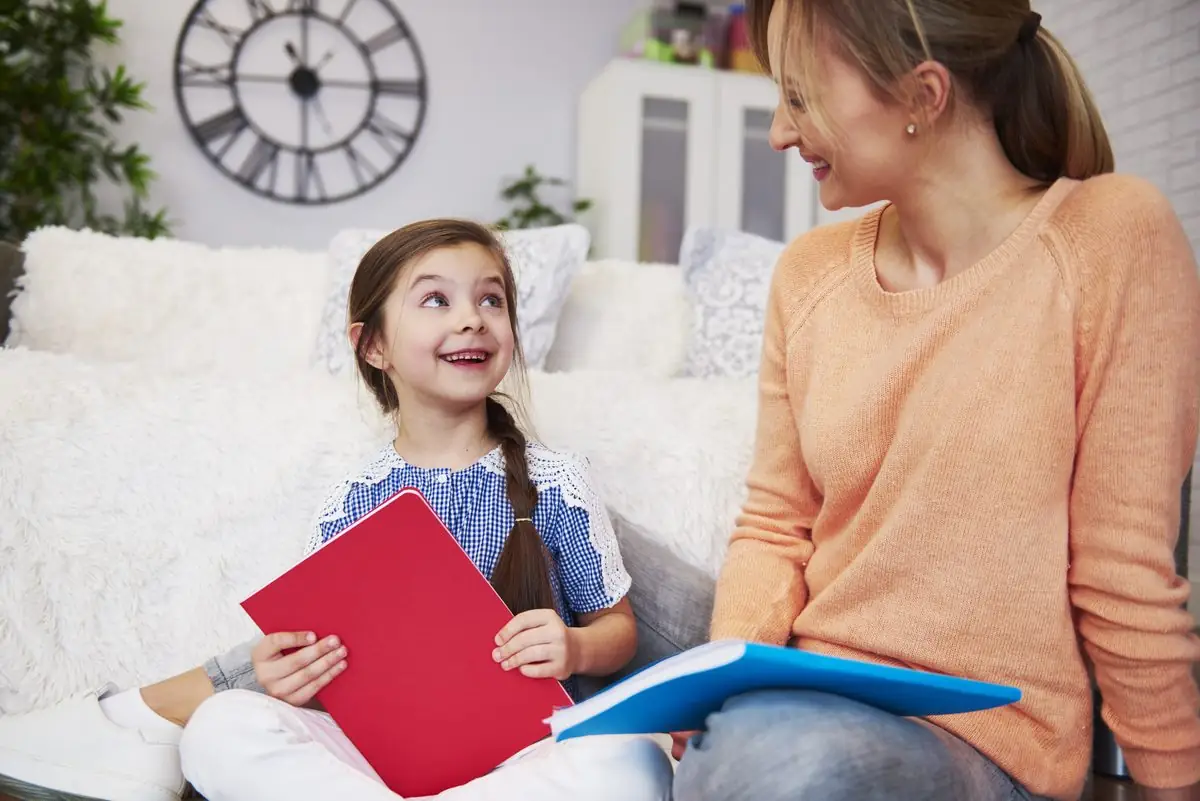 A mother and her young daughter smile at each other over school workbooks on a sofa