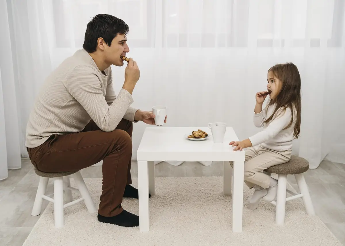 A father and his young daughter share a snack at a small white table at home