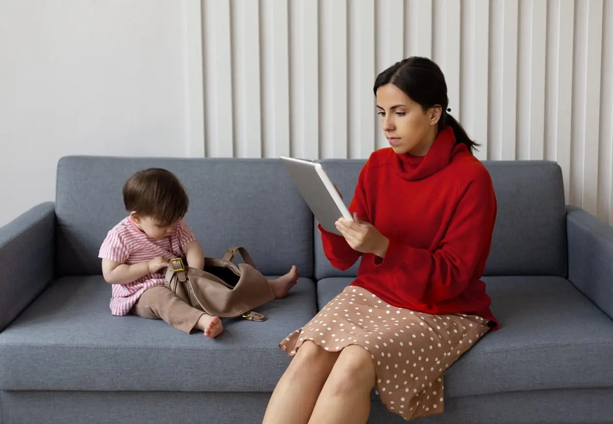 A mother reads from a tablet on a grey sofa while her toddler plays beside her
