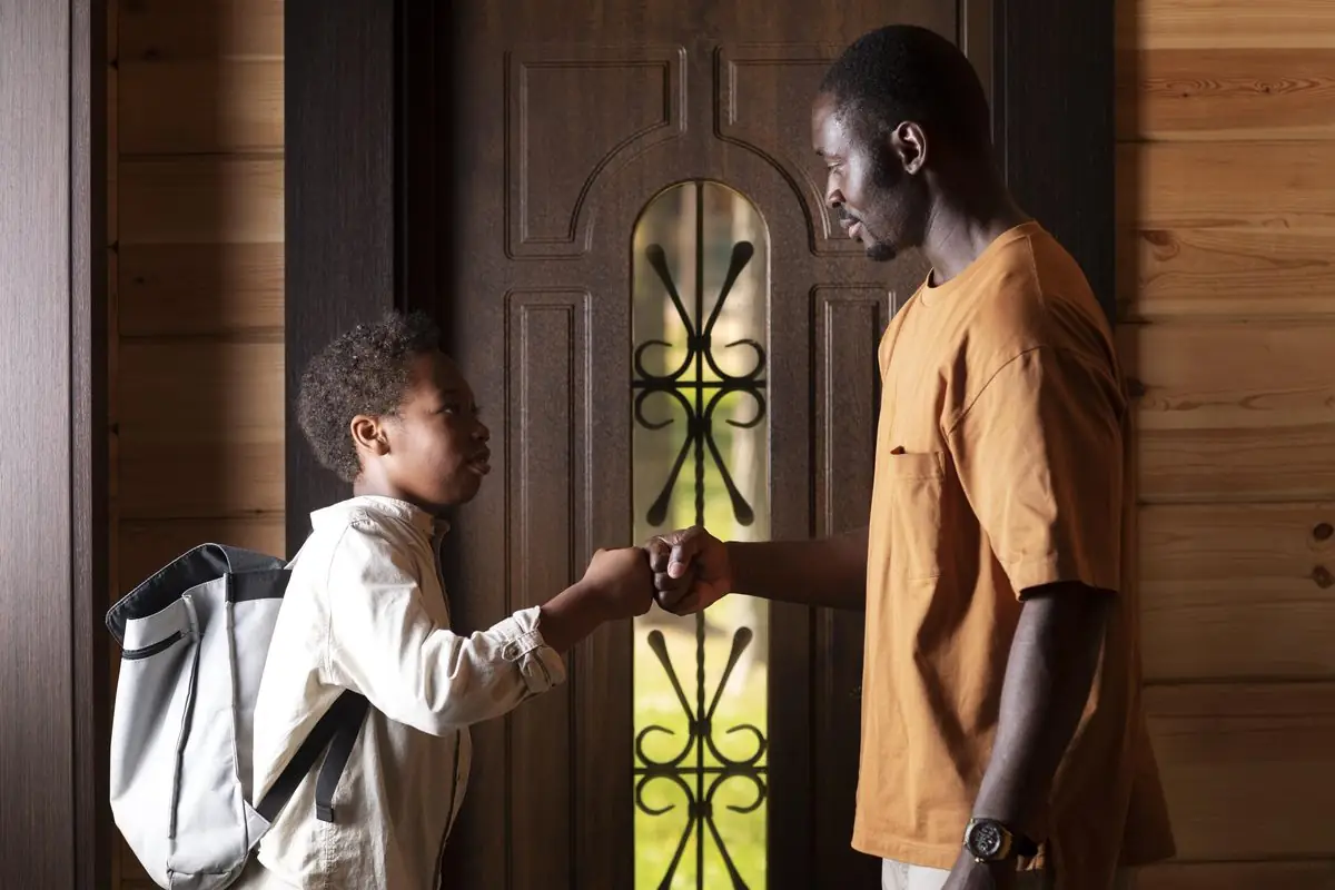A man in an orange t-shirt fist-bumps a boy with a backpack in a wooden doorway