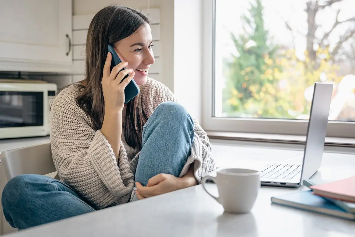 A young woman on the phone at her kitchen table with a laptop open