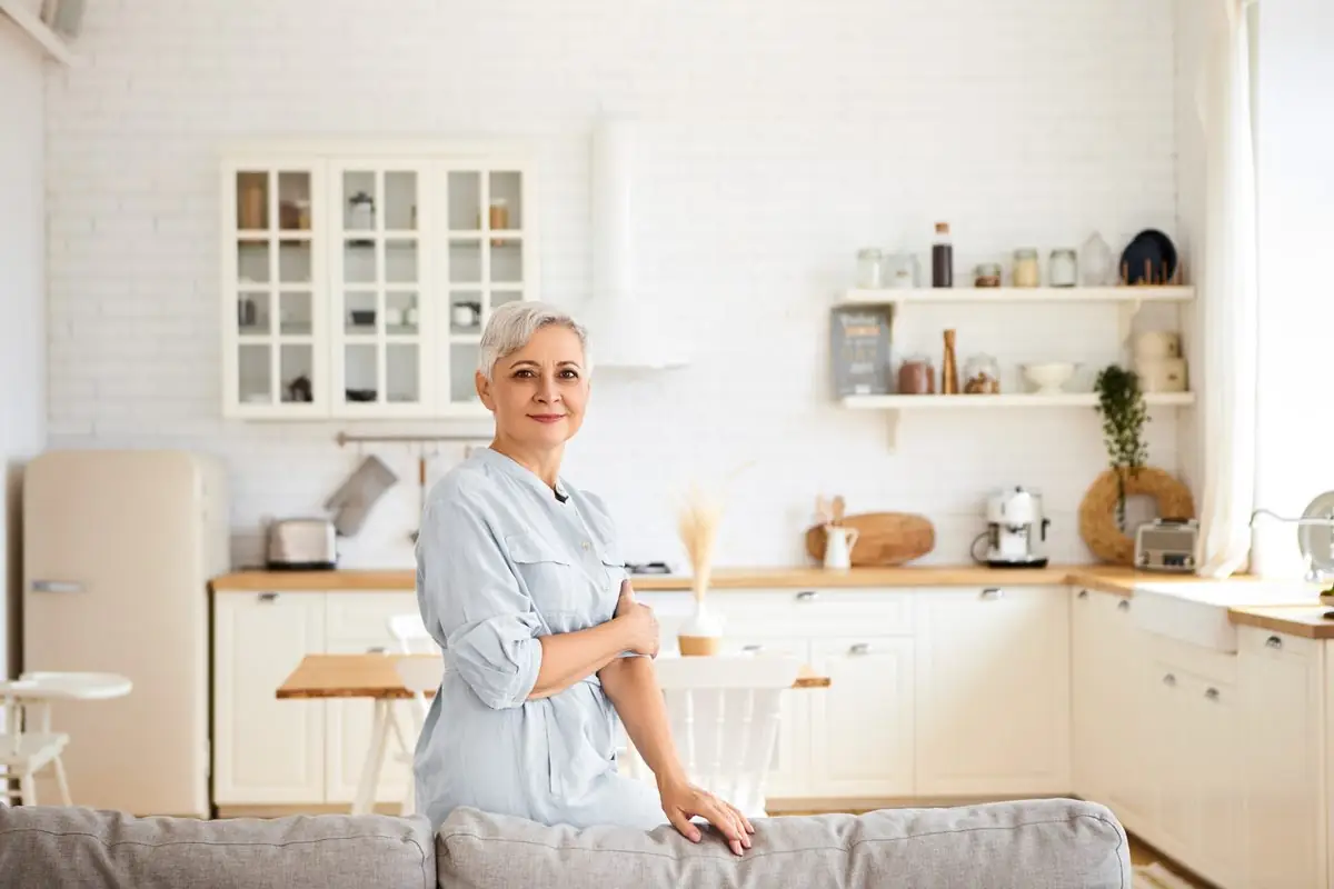 A woman standing in her bright living room with the kitchen behind her