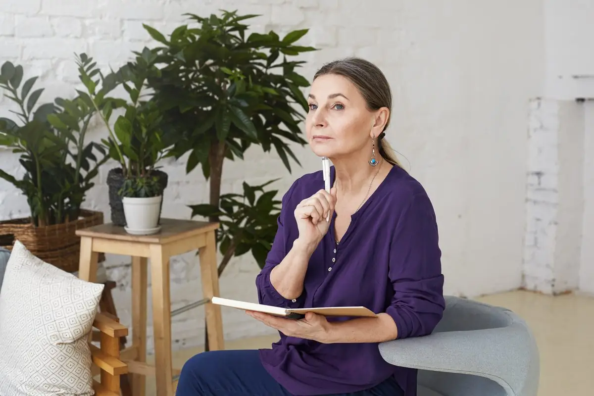 A mature woman sitting in an armchair holding a notebook and looking thoughtful