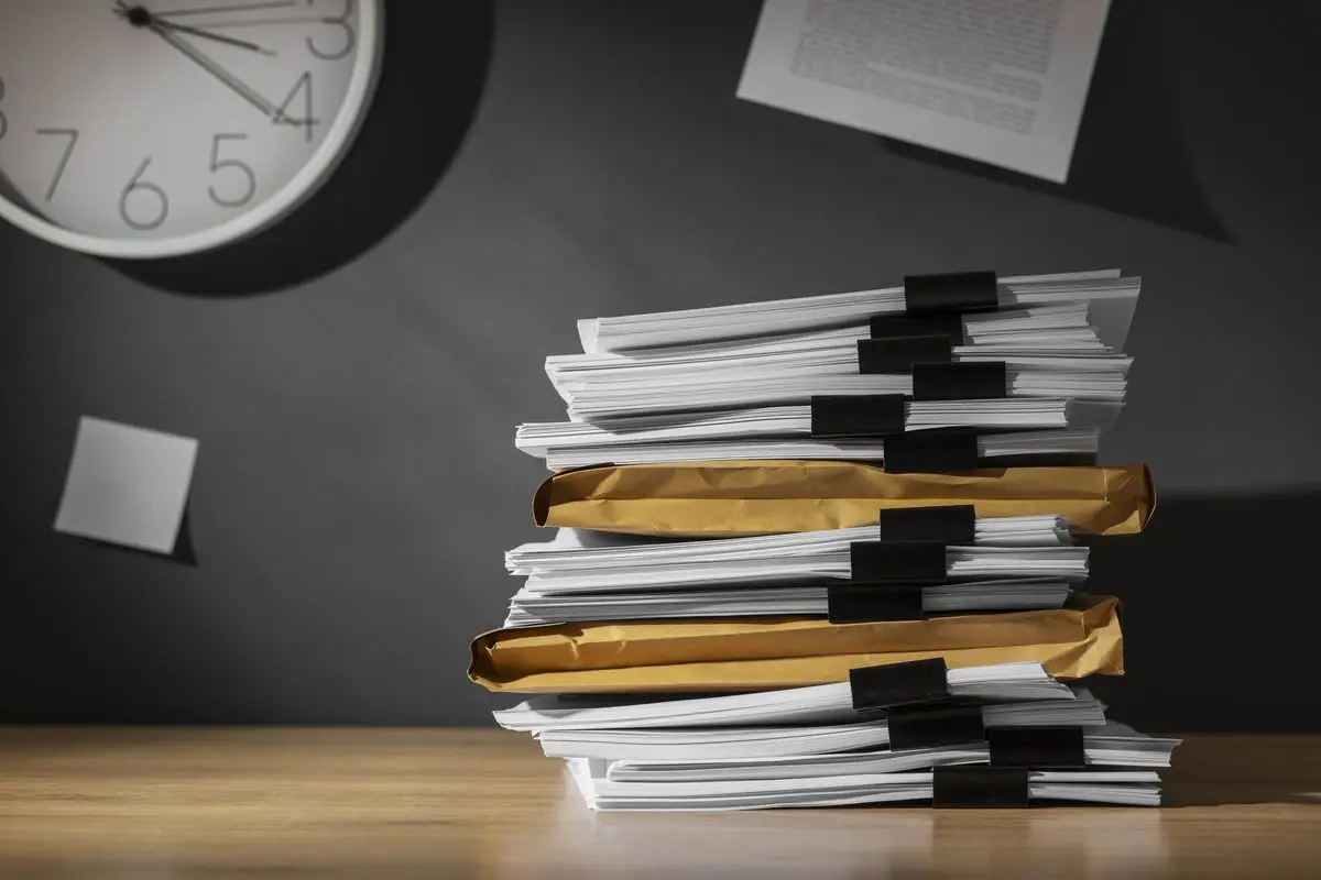 Tall stacks of documents and envelopes on a desk beneath a wall clock