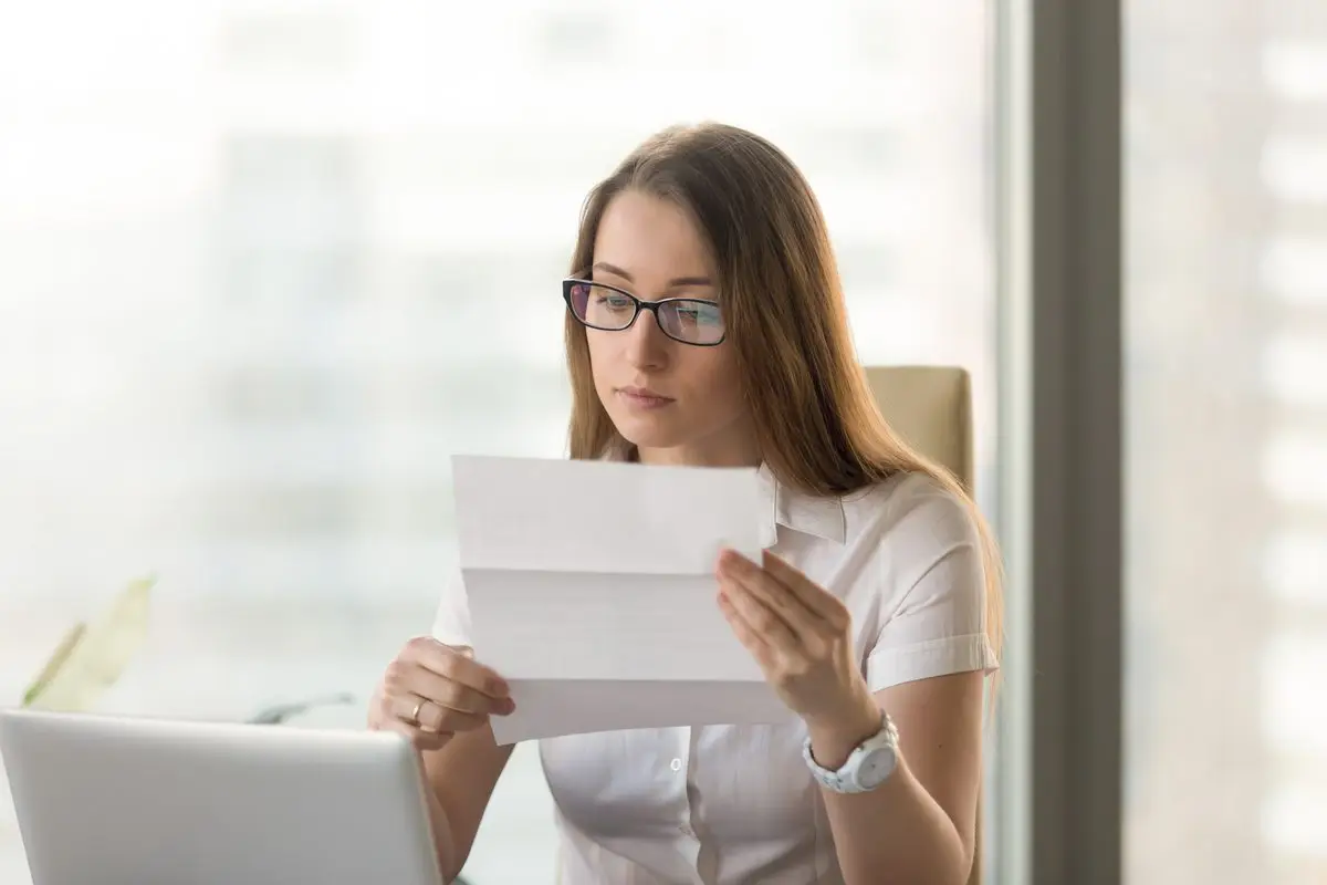 A woman reading through a document at her desk beside an open laptop
