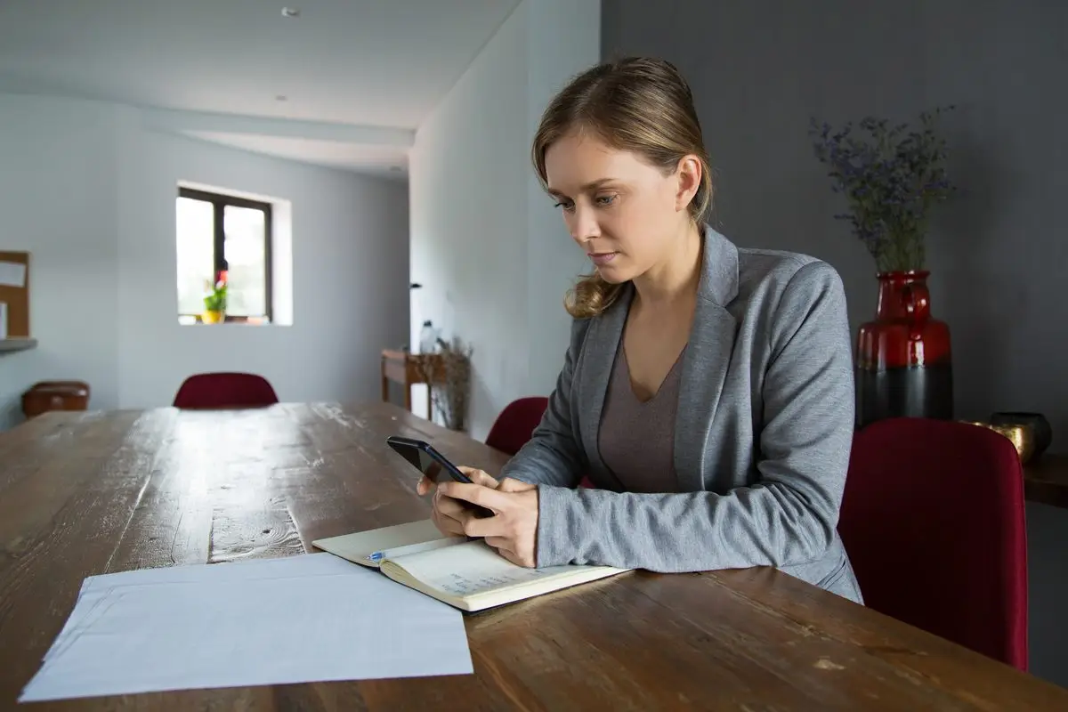 A woman at a long wooden table with a notebook, loose papers and a phone, working through terms by herself