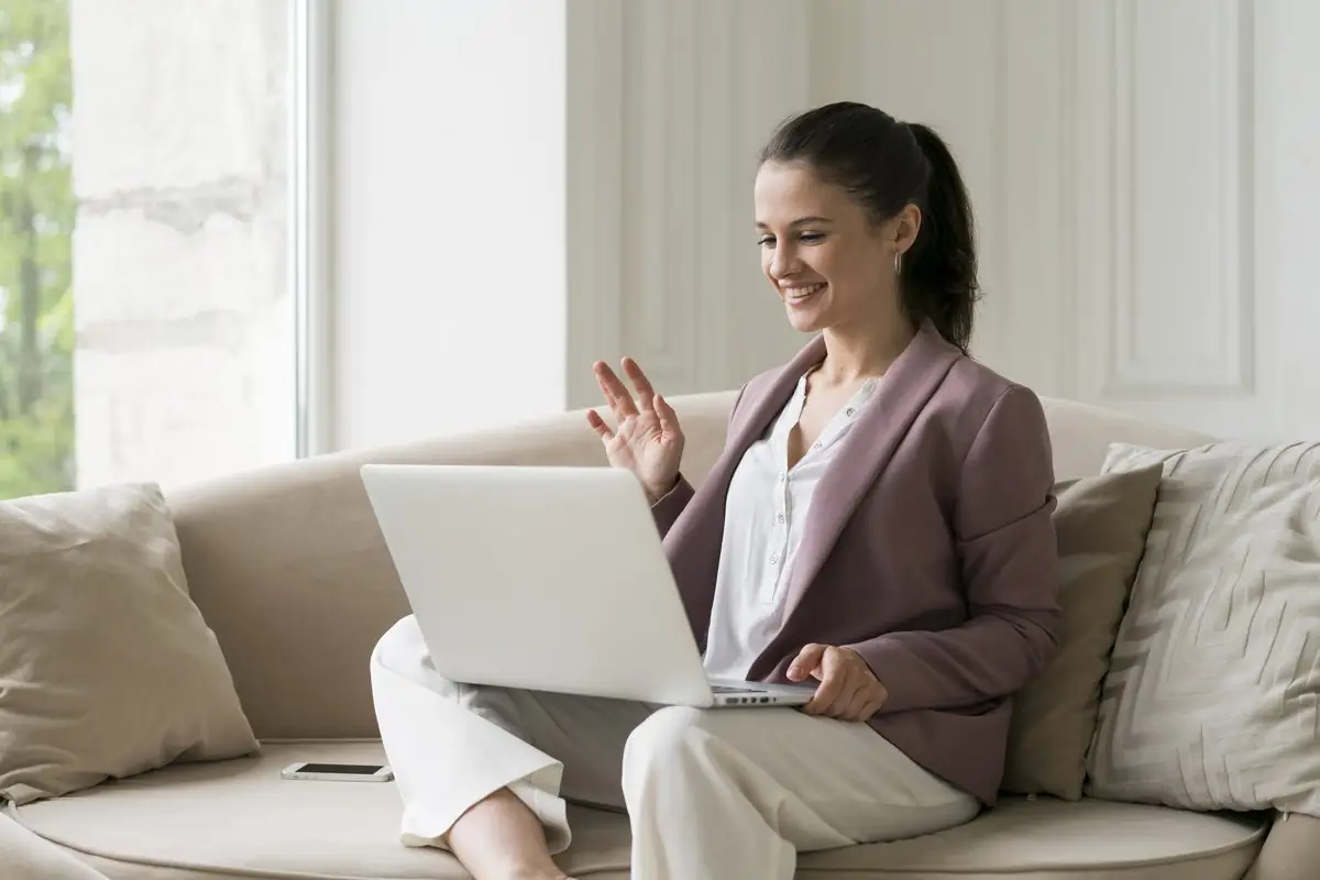 A smiling woman on a sofa waving to her laptop during a video consultation