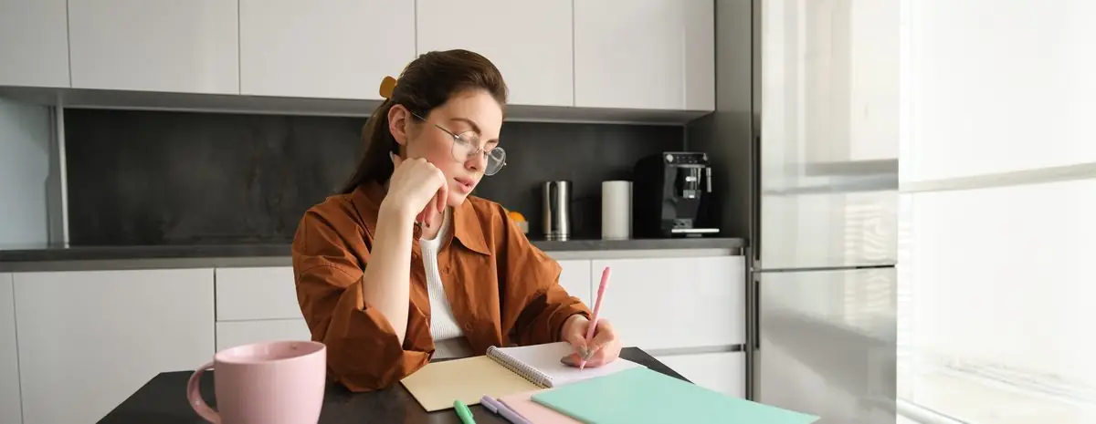 A woman at a kitchen table writing questions in a notebook beside a mug
