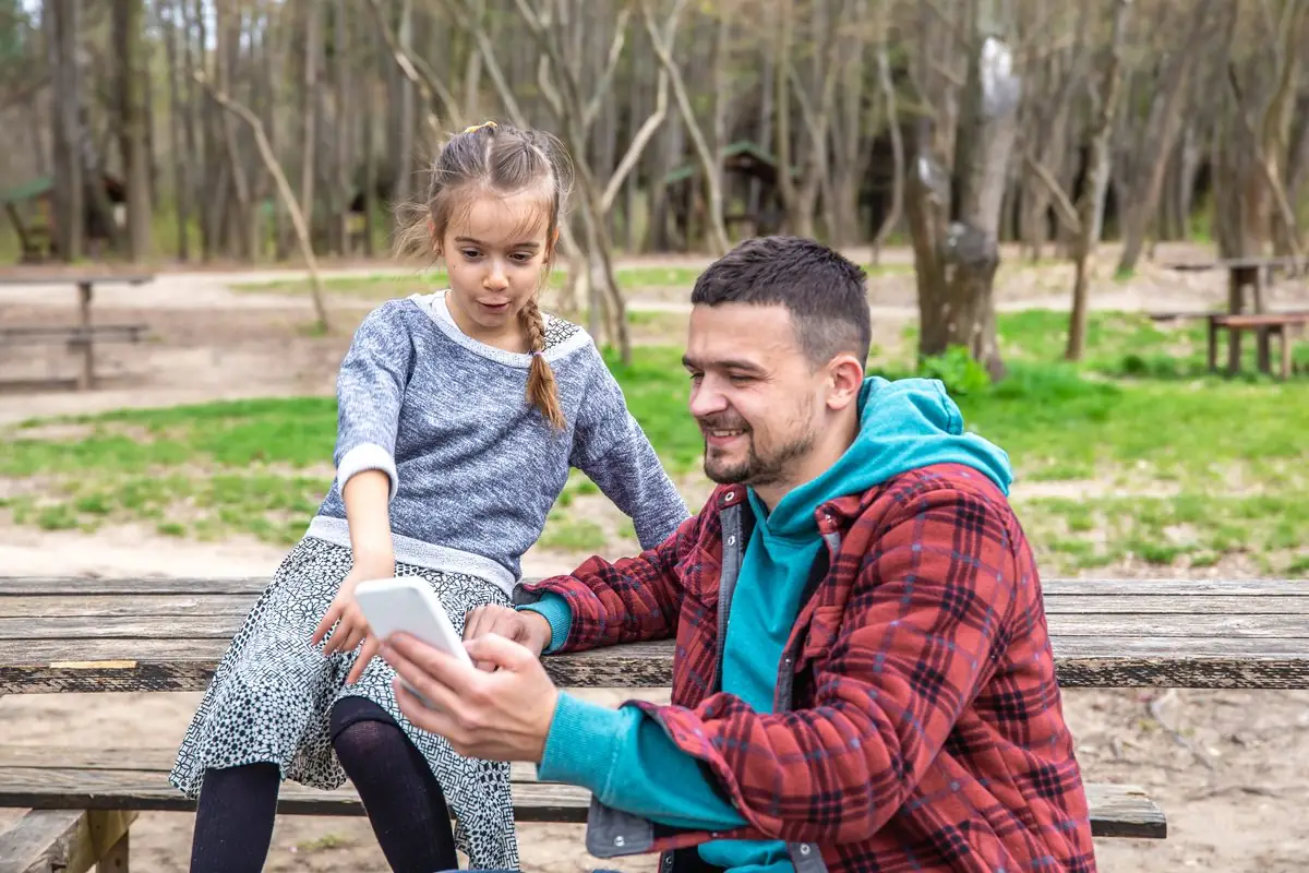 A father and his daughter sharing a phone at a picnic table in a park