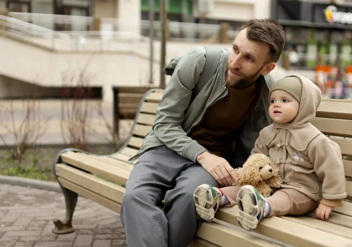 A father sitting on a bench with his toddler daughter and her teddy bear