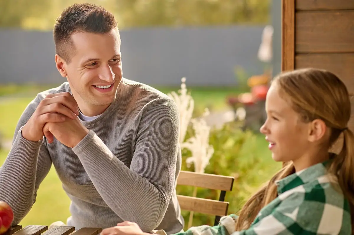 A father smiling and listening closely as his daughter talks at an outdoor table