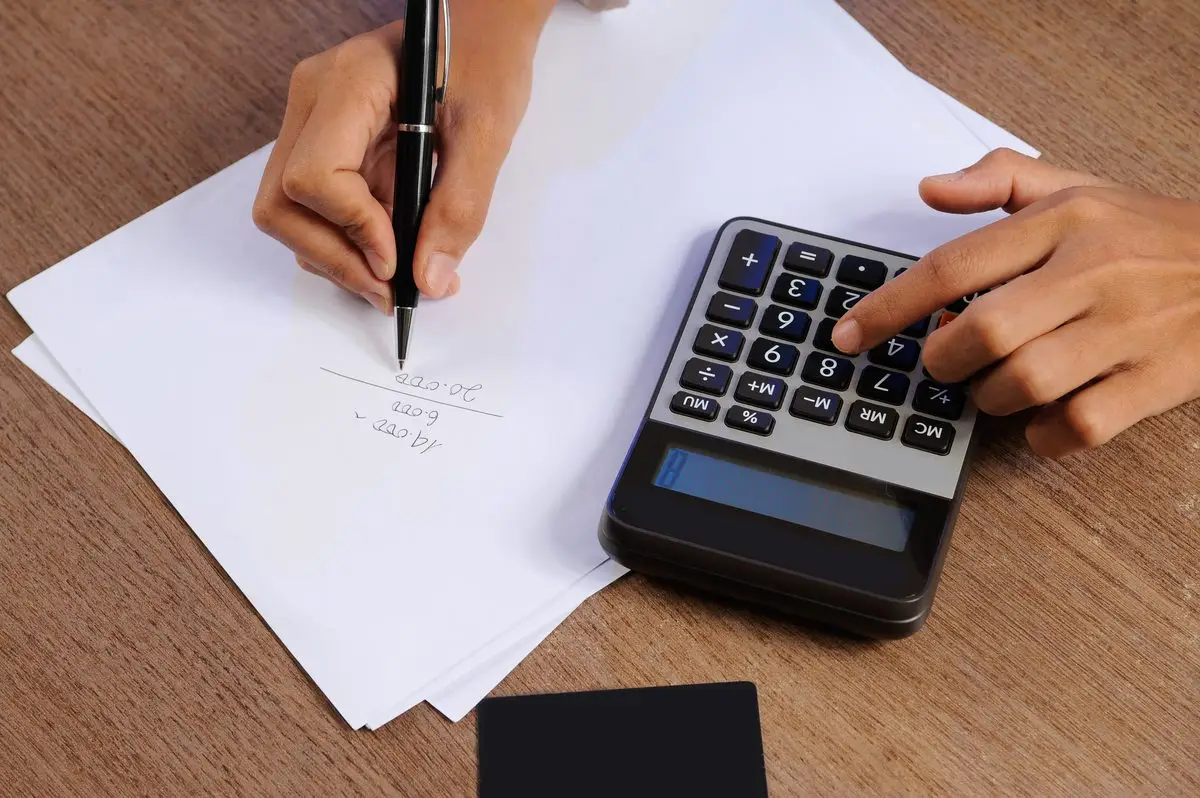 A hand writing figures on paper next to a calculator
