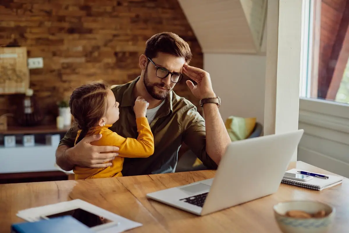 A parent at a laptop at home reading a worrying message while holding a young child