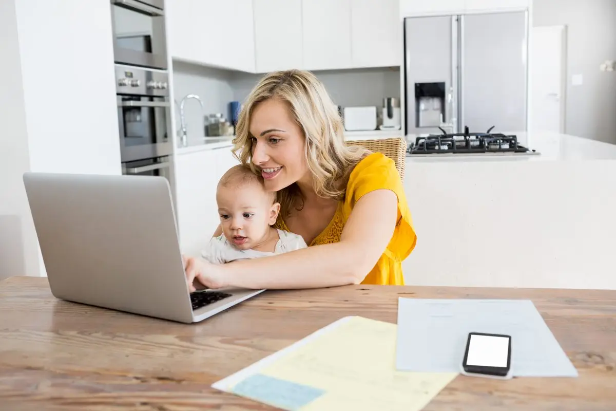 A mother at the kitchen table going over papers on a laptop with her baby on her lap